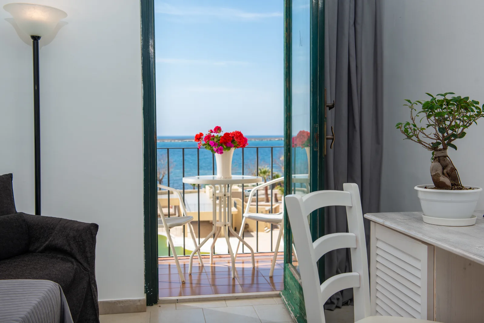 Balcone di una camera affacciato sul mare con tavolino, sedie bianche e vaso di fiori rossi