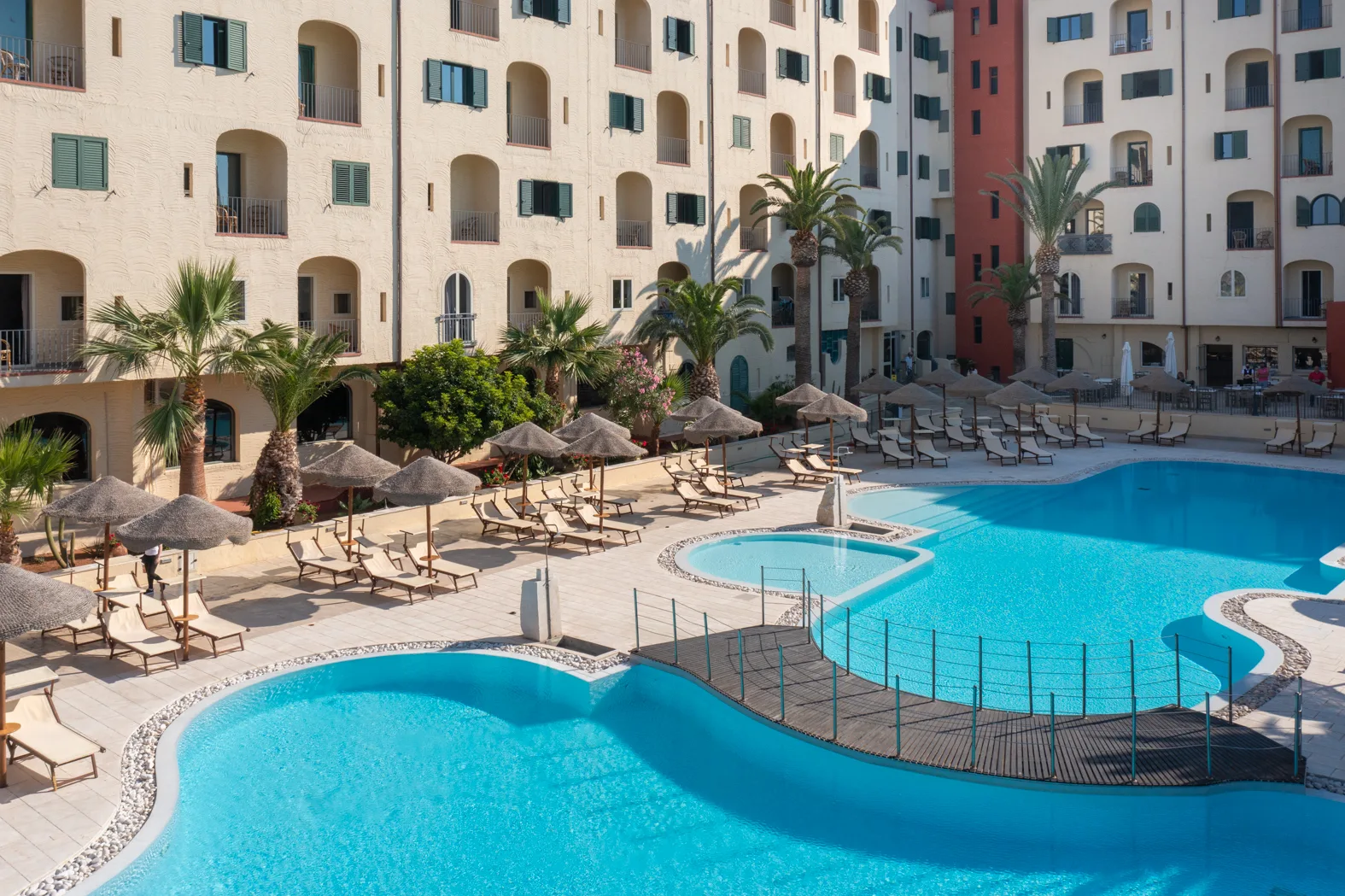 Piscine dell'hotel con ombrelloni in paglia, lettini, palme e passerella in legno tra le vasche