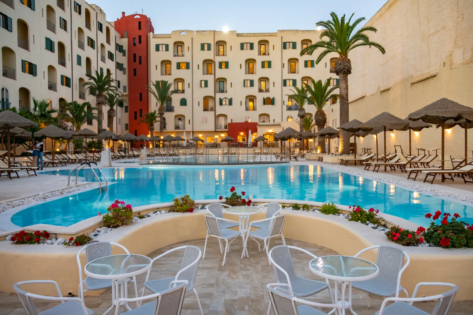 Area piscina al tramonto con tavolini bianchi, fiori rossi, ombrelloni illuminati e edificio dell'hotel sullo sfondo