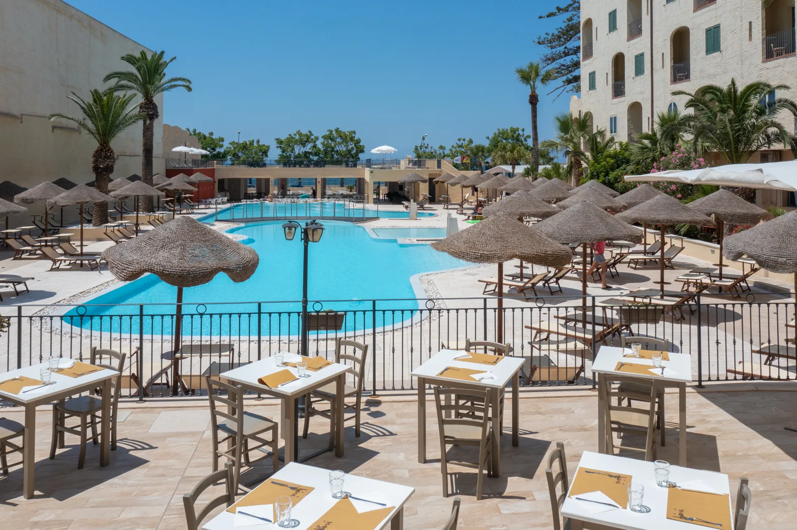 Terrazza ristorante con tavoli apparecchiati affacciata sulla piscina con ombrelloni in paglia e palme