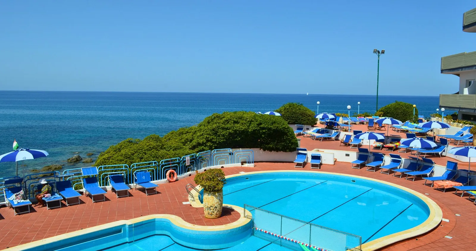 Piscina all'aperto con lettini e ombrelloni blu su terrazza affacciata sul mare azzurro