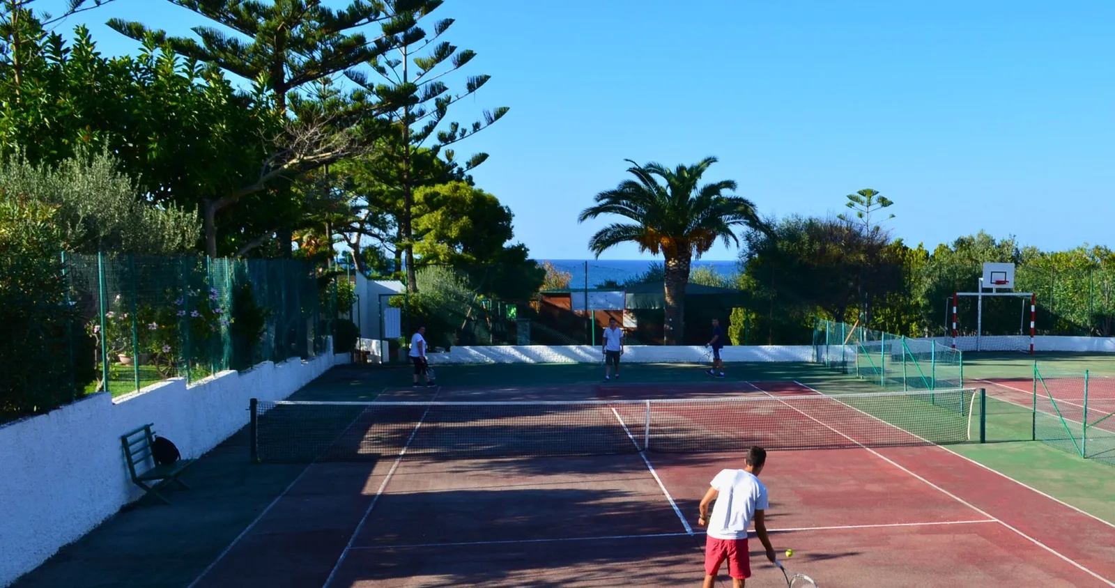 Campo da tennis con giocatori circondato da palme e alberi con vista sul mare