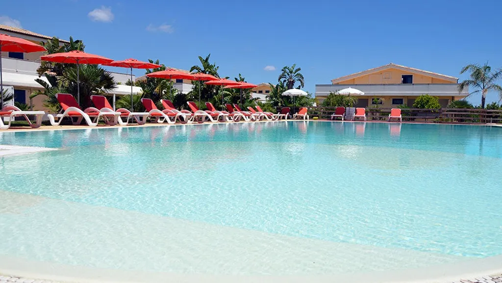 Piscina con acqua cristallina, lettini con ombrelloni rossi ed edifici dell'hotel